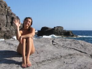woman sitting in a bikini on a rock by the sea waving